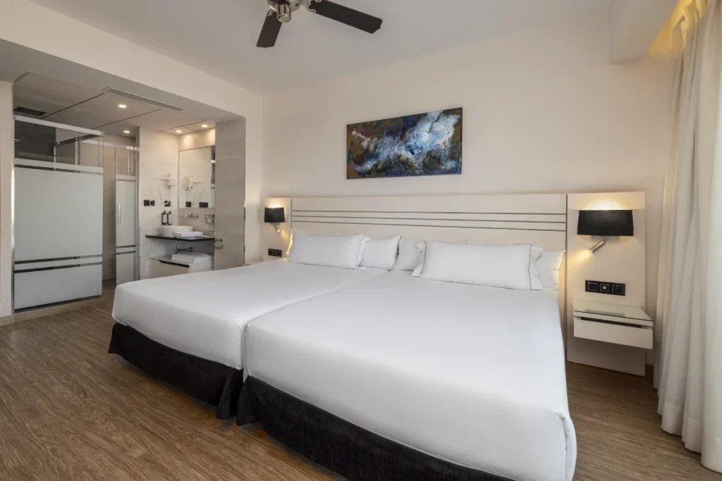 Dormitorio moderno con cama blanca, iluminaci&oacute;n y ba&ntilde;o integrado al fondo