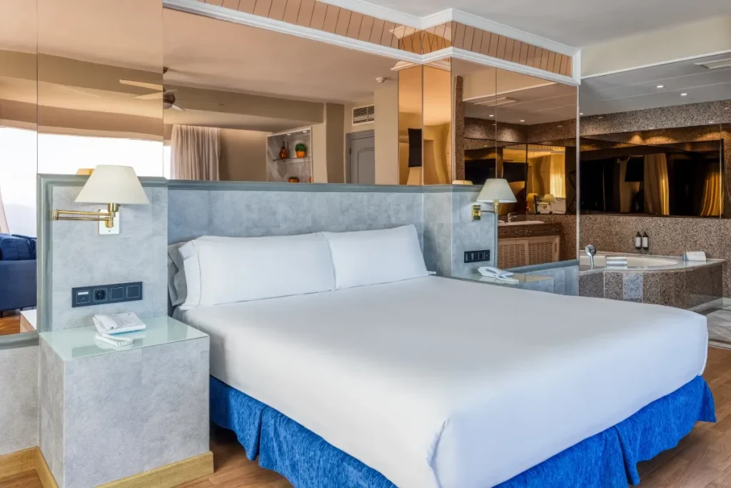 Habitaci&oacute;n de hotel moderna con cama blanca, sof&aacute; azul y ba&ntilde;o visible.