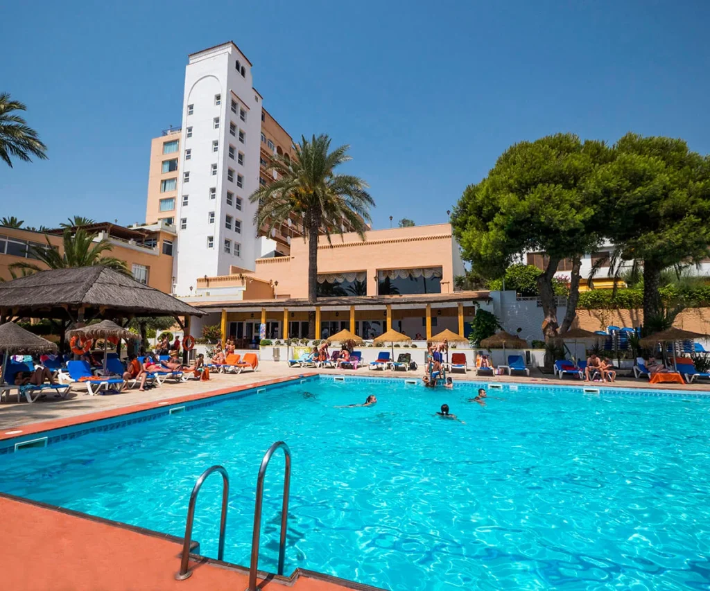 Piscina de hotel con gente y edificios blancos, palmeras y tumbonas azules