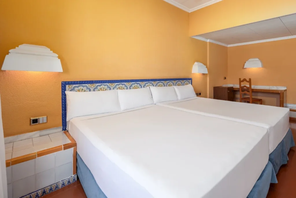 Dormitorio con cama doble blanca, l&aacute;mparas y escritorio, paredes naranjas