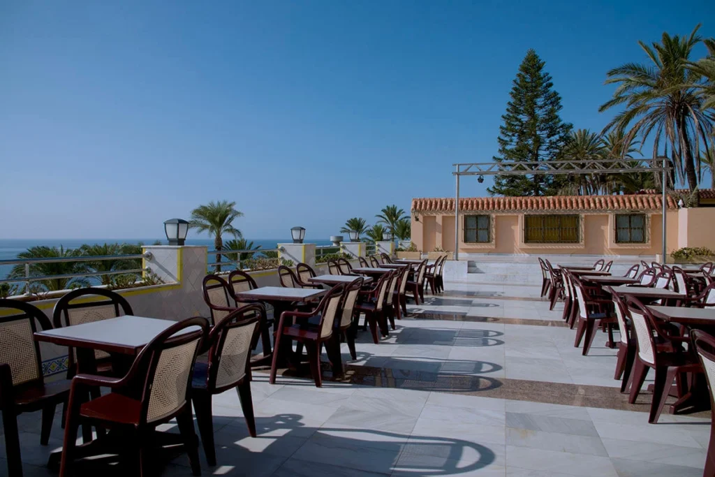 Terraza con mesas y sillas junto al mar, palmeras y cielo despejado.
