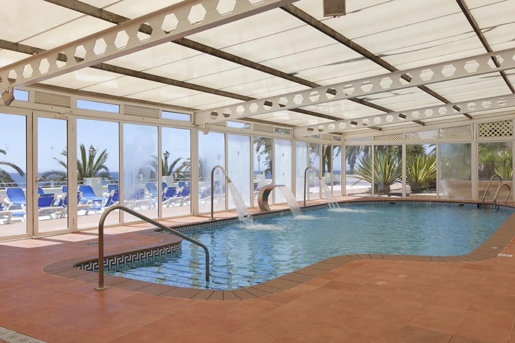 Piscina cubierta con techo de cristal, vista al mar y tumbonas al fondo