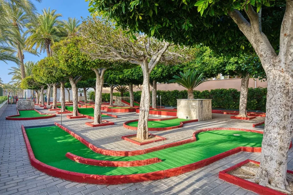 Jard&iacute;n con senderos y c&eacute;sped verde con bordes rojos entre palmeras y &aacute;rboles.