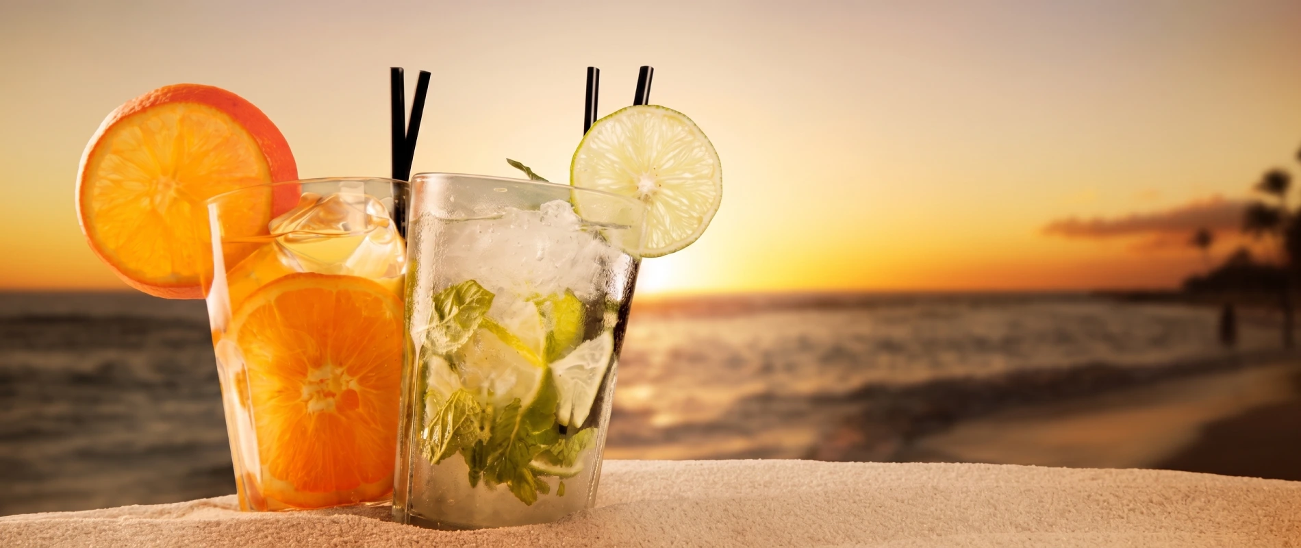 C&oacute;cteles con hielo y rodajas de naranja y lim&oacute;n en la playa al atardecer