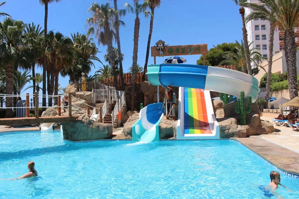 Parque acuático con tobogán azul y piscina en un entorno tropical con palmeras