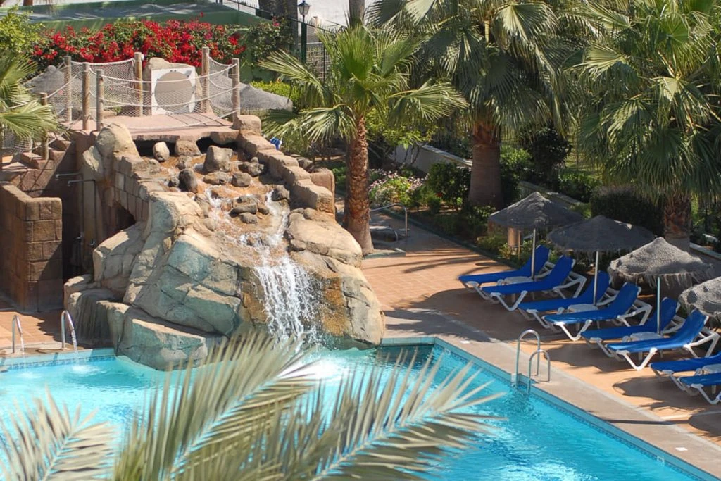 Piscina con cascada, tumbonas azules y palmeras en un jardín tropical