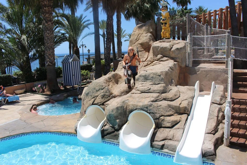 Piscina con tobogán blanco, rocas, palmeras y figura junto al mar.