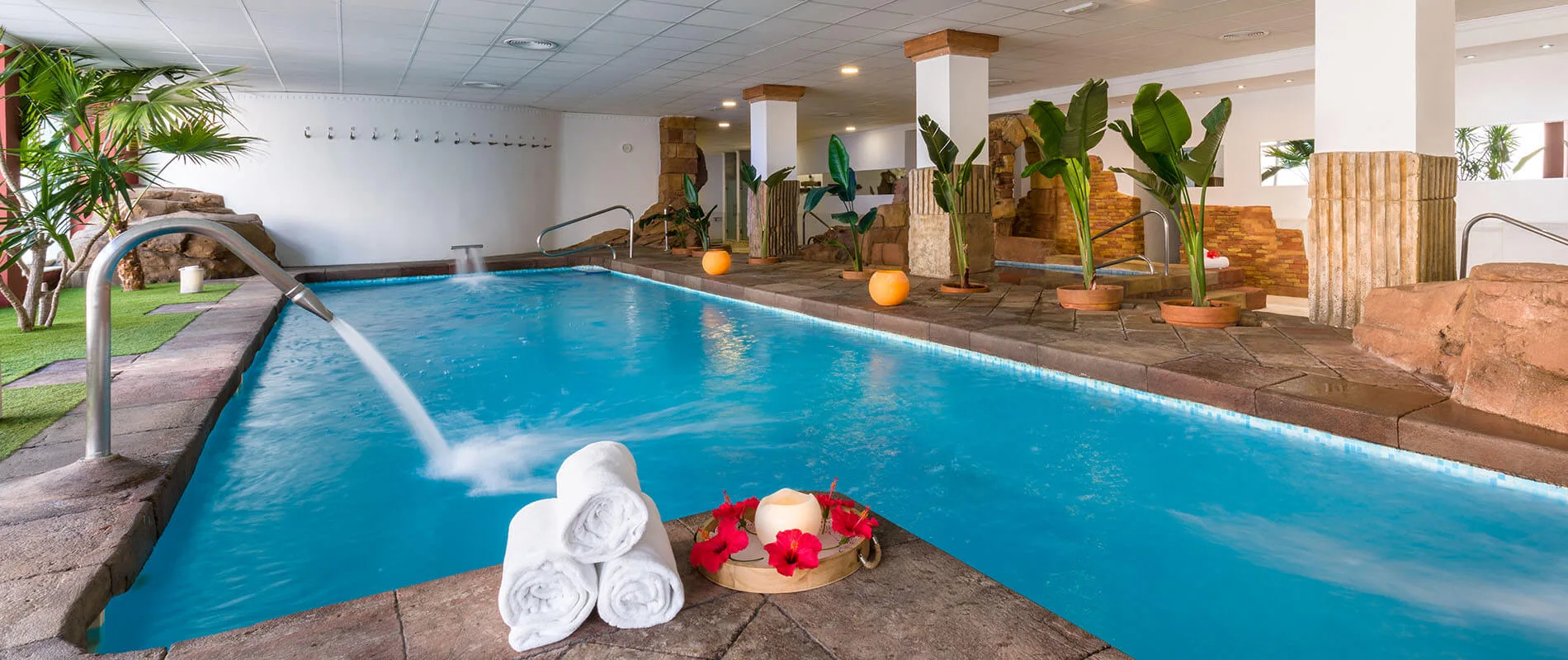 Piscina interior con plantas tropicales, chorros de agua y toallas enrolladas