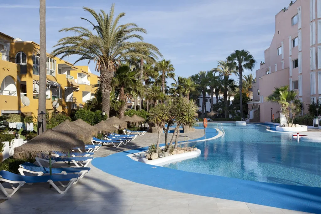 Piscina azul con palmeras y tumbonas junto a edificios blancos y amarillos