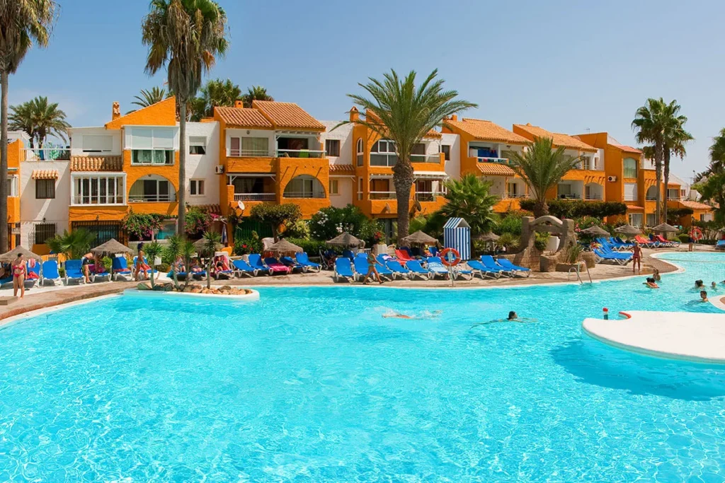 Piscina azul con tumbonas y villas mediterr&aacute;neas con palmeras al fondo