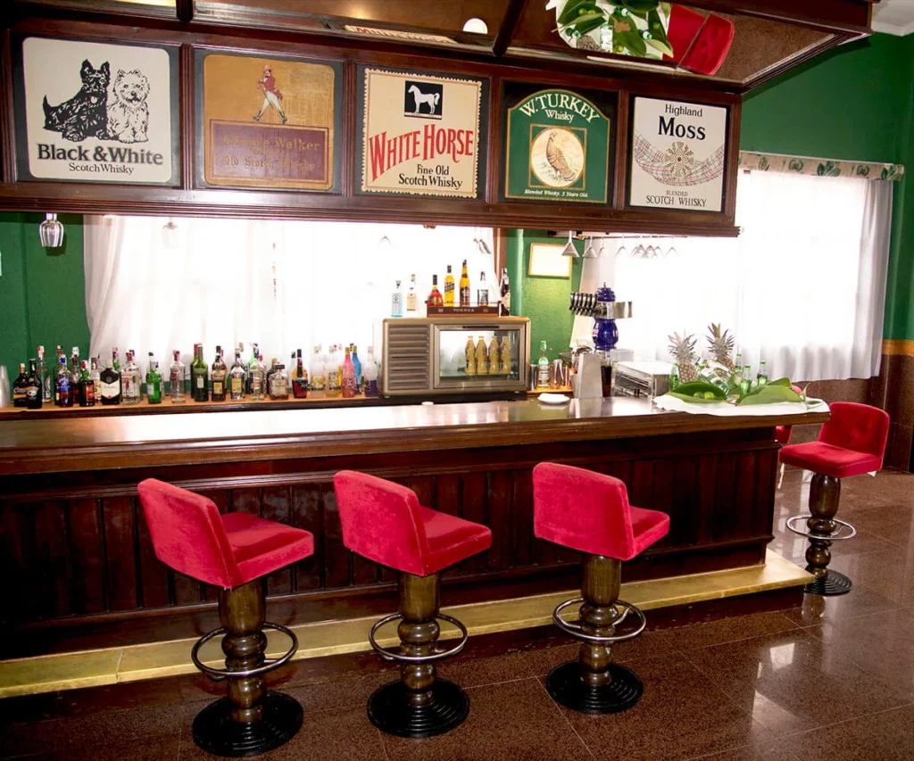 Bar con botellas de whisky, cartelera y taburetes rojos en un interior.