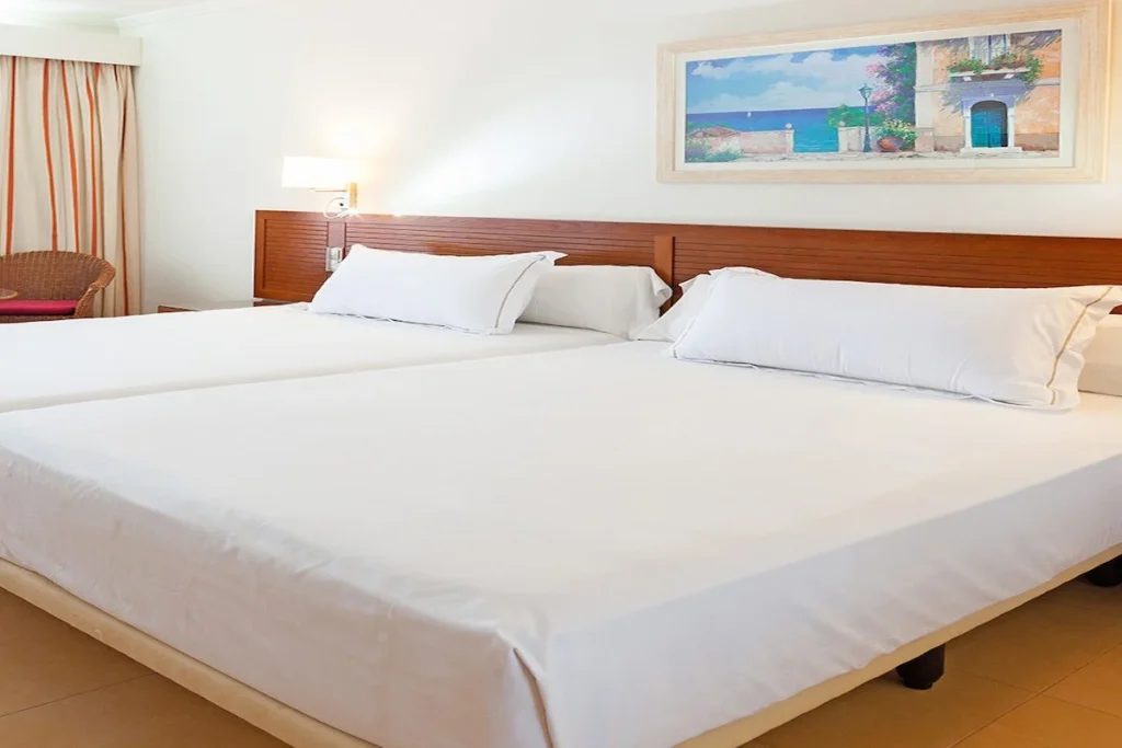 Cama doble con s&aacute;banas blancas y cuadro decorativo en habitaci&oacute;n hotelera