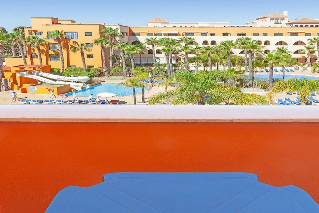 Hotel con piscina y palmeras, vista desde balc&oacute;n en d&iacute;a soleado.