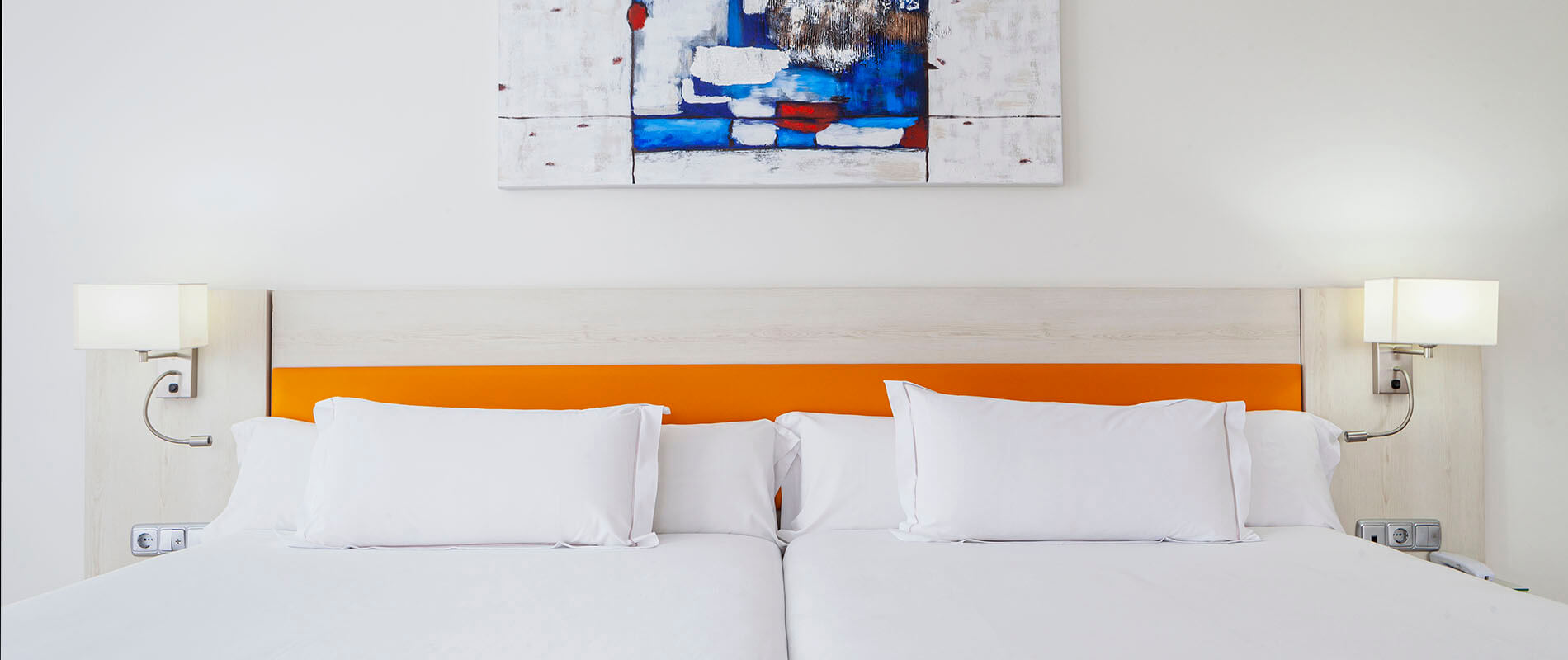Cama doble con cojines blancos y cabecero naranja, l&aacute;mparas y cuadros abstractos.