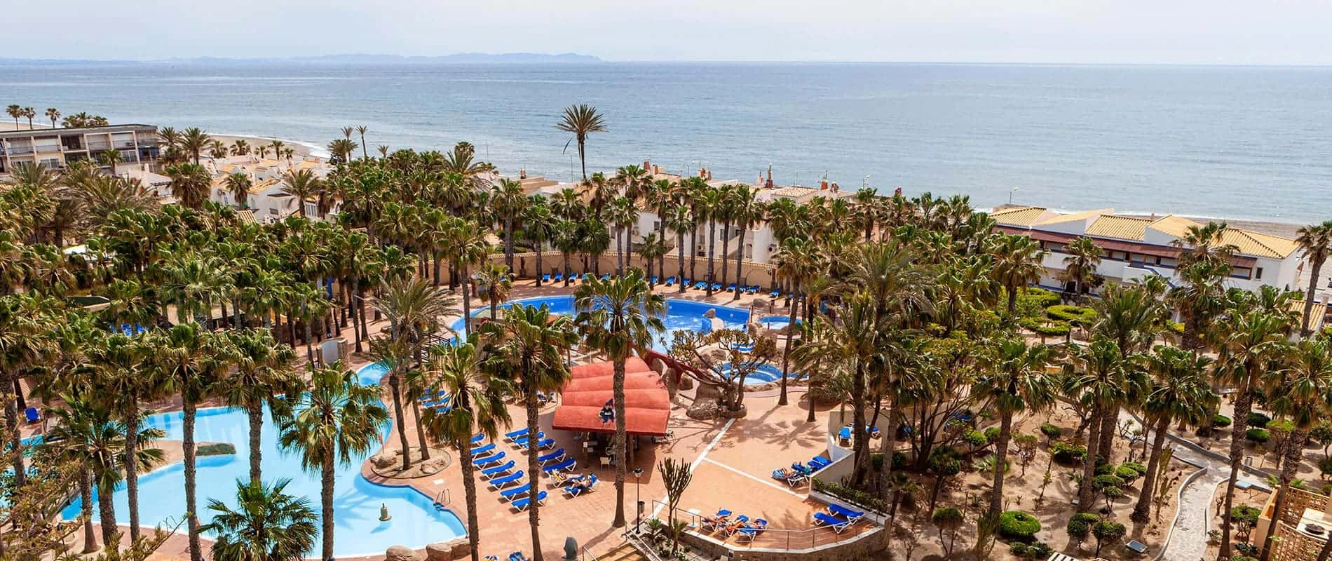 Hotel junto al mar, palmeras y piscinas azules con tumbonas, vista panor&aacute;mica