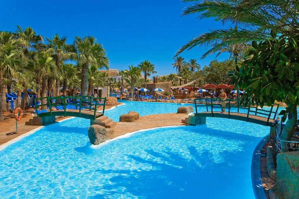 Piscina de hotel con tumbonas y sombrillas, rodeada de palmeras y cielo azul.