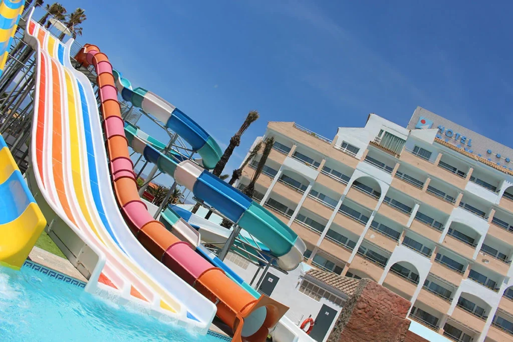 Toboganes de colores y hotel junto a piscina, con cielo azul.
