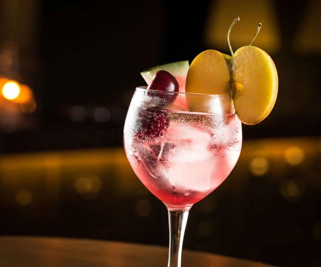 C&oacute;ctel rosado con hielo y cereza, naranja y lim&oacute;n en copa al fondo oscuro