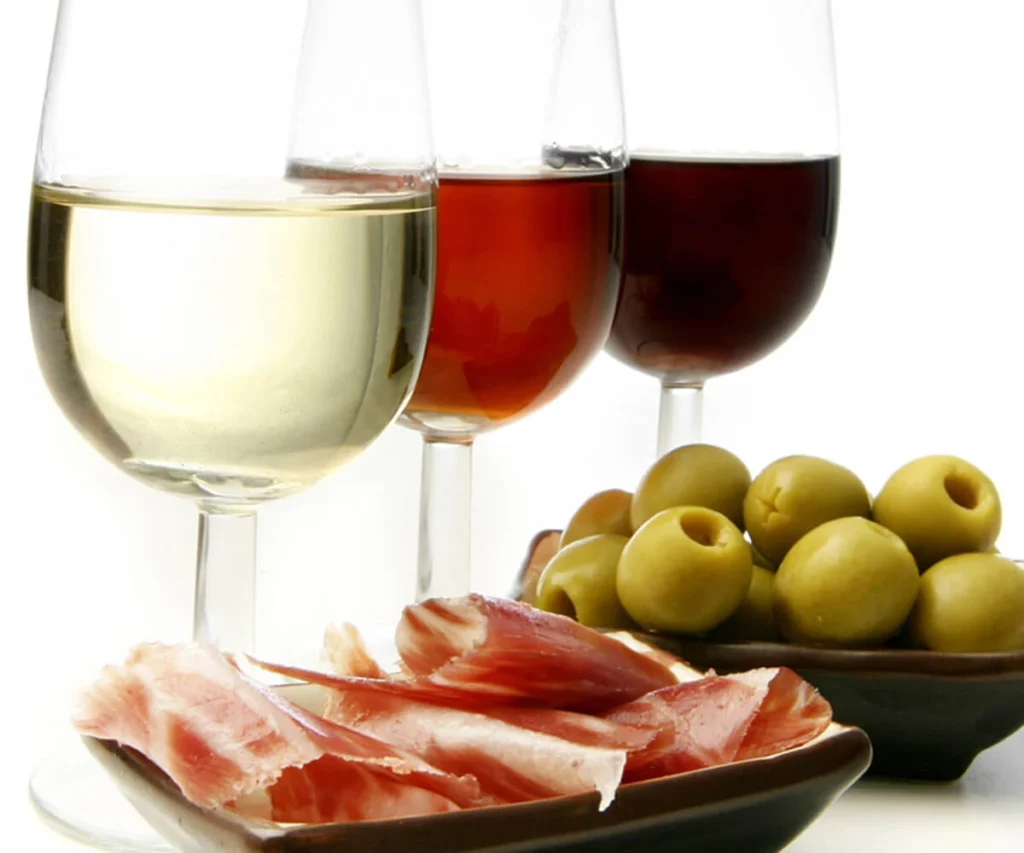 Tres copas con vino, jam&oacute;n y aceitunas sobre una mesa blanca.