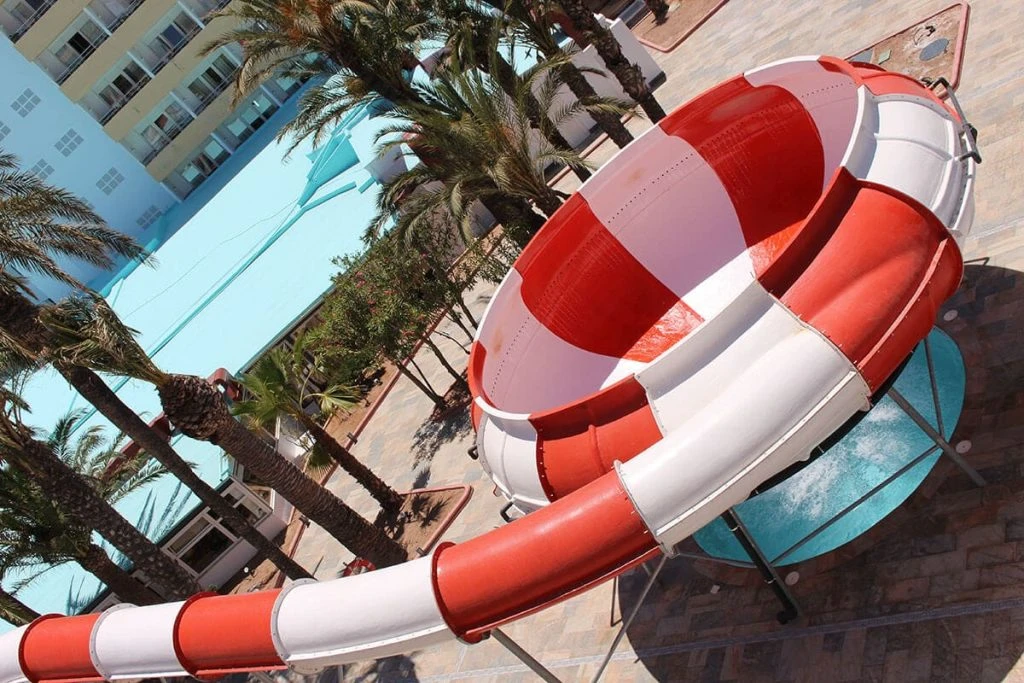 Tobogán rojo y blanco en un resort con palmeras y piscina azul.