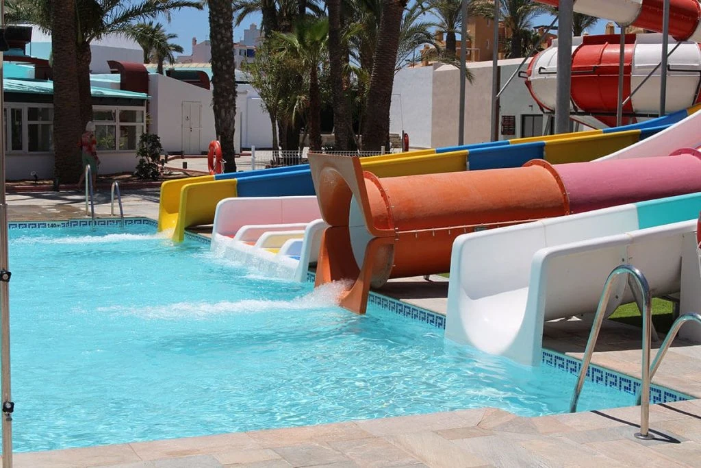 Toboganes de colores junto a una piscina con palmeras y edificios al fondo.