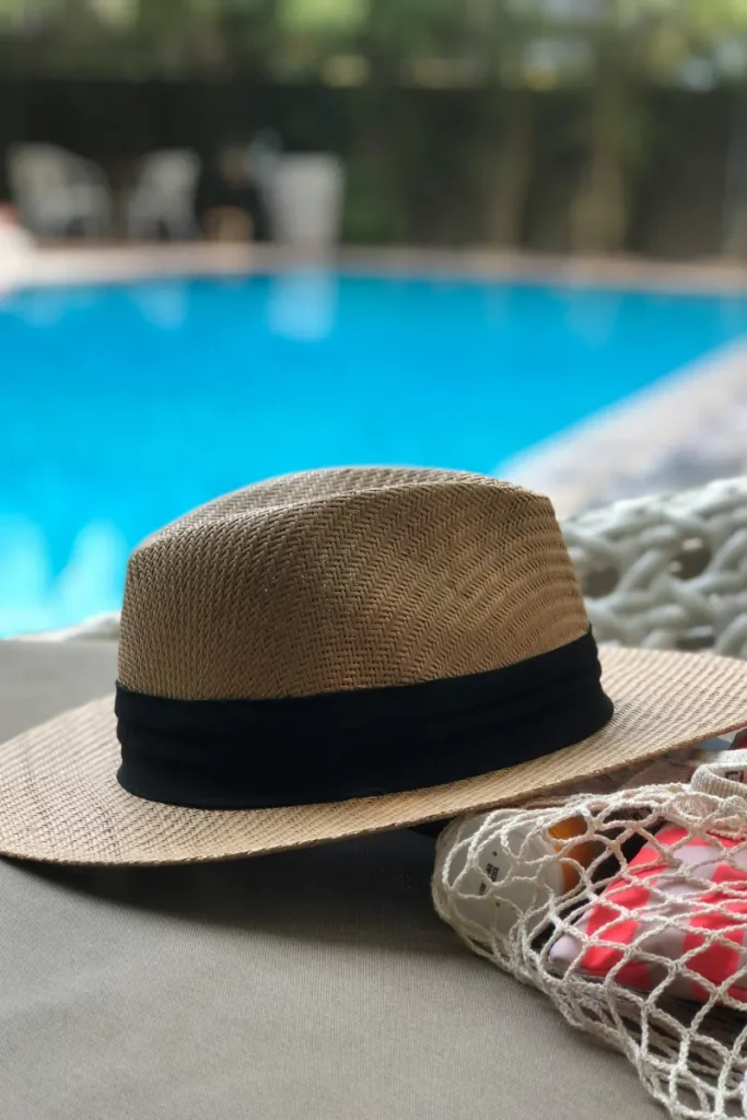 Sombrero de paja beige sobre coj&iacute;n junto a una piscina azul desenfocada