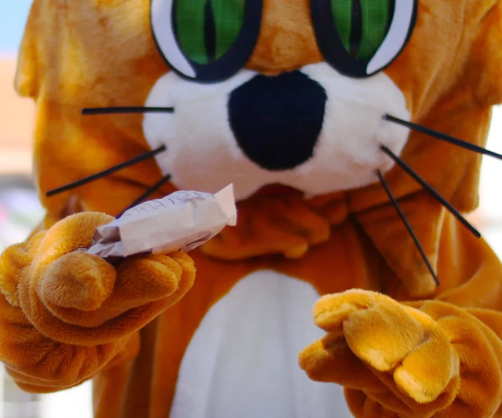 Mu&ntilde;eco de peluche naranja con ojos verdes y sonrisa, sosteniendo una bolsita