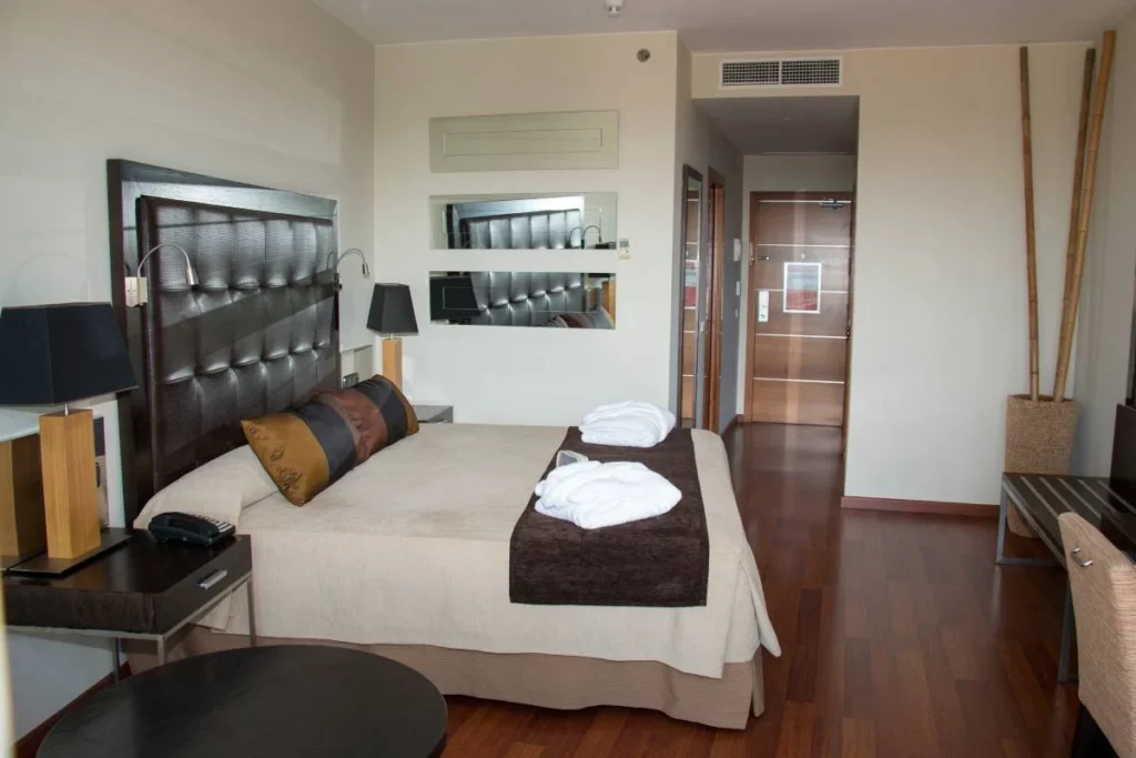 Habitaci&oacute;n de hotel moderna con cama, mesitas y puerta de entrada al fondo