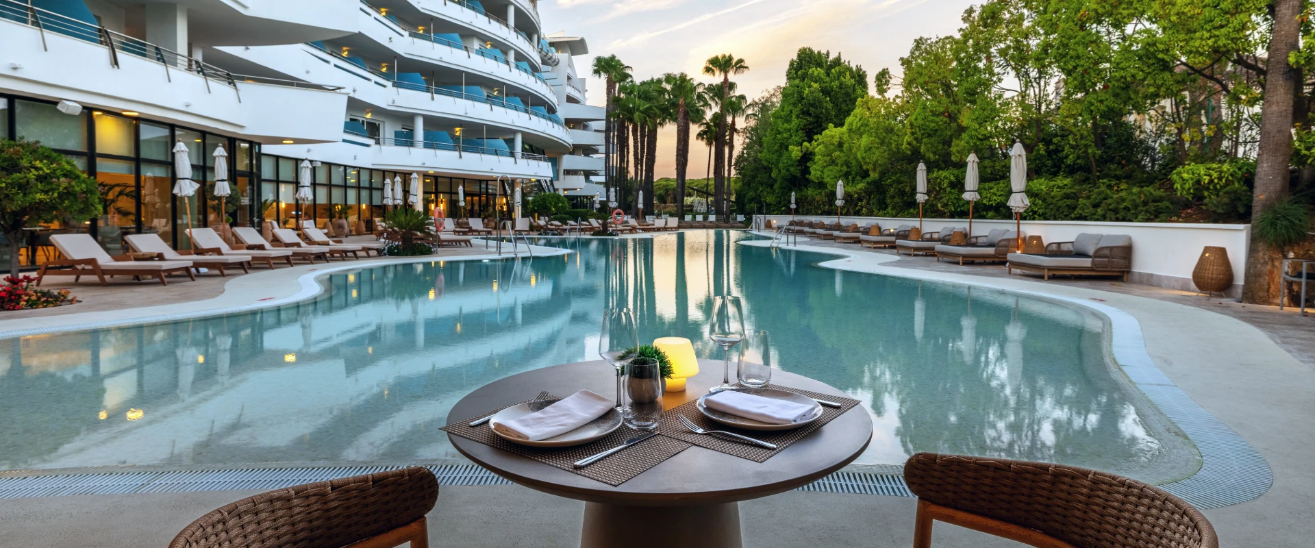 Piscina de hotel con mesa servida y tumbonas, rodeada de palmeras y jardines