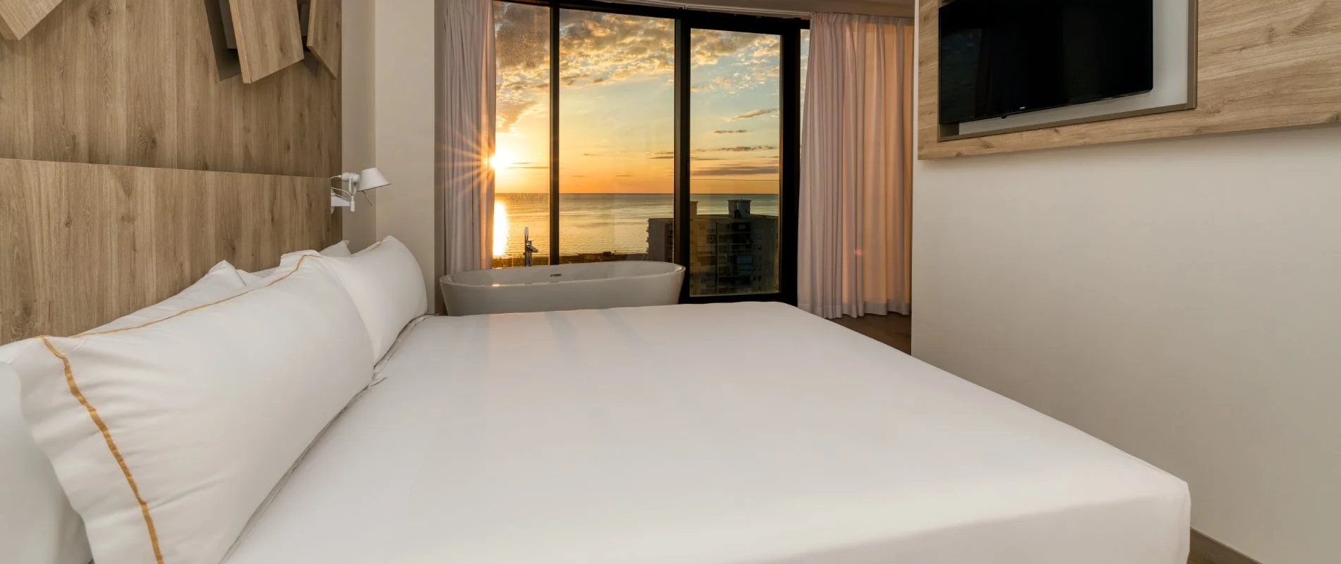 Dormitorio con cama blanca y ba&ntilde;era frente a ventanales al atardecer en la costa