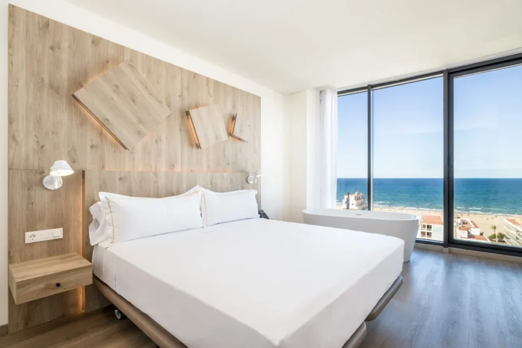Dormitorio moderno con cama blanca y ventana al mar, luz natural.