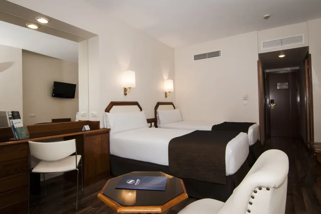 Habitaci&oacute;n de hotel moderna con camas, escritorio, sillones y TV.