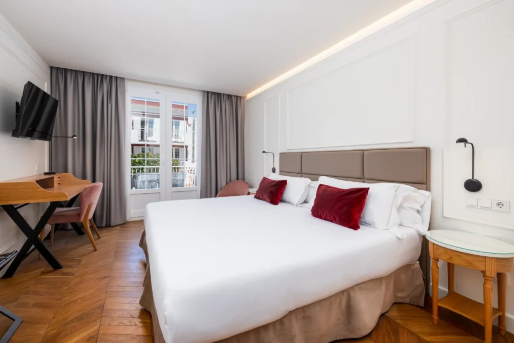 Dormitorio moderno con cama blanca, cojines rojos y escritorio con TV.