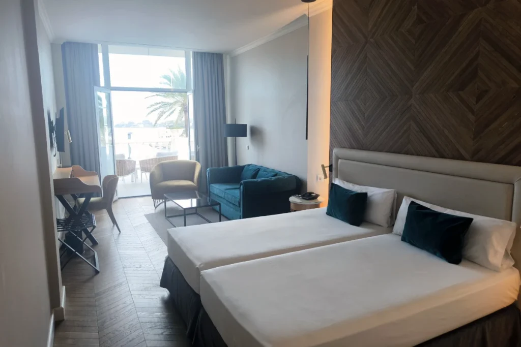 Dormitorio con cama doble, sof&aacute; azul y salida a terraza con palmeras.