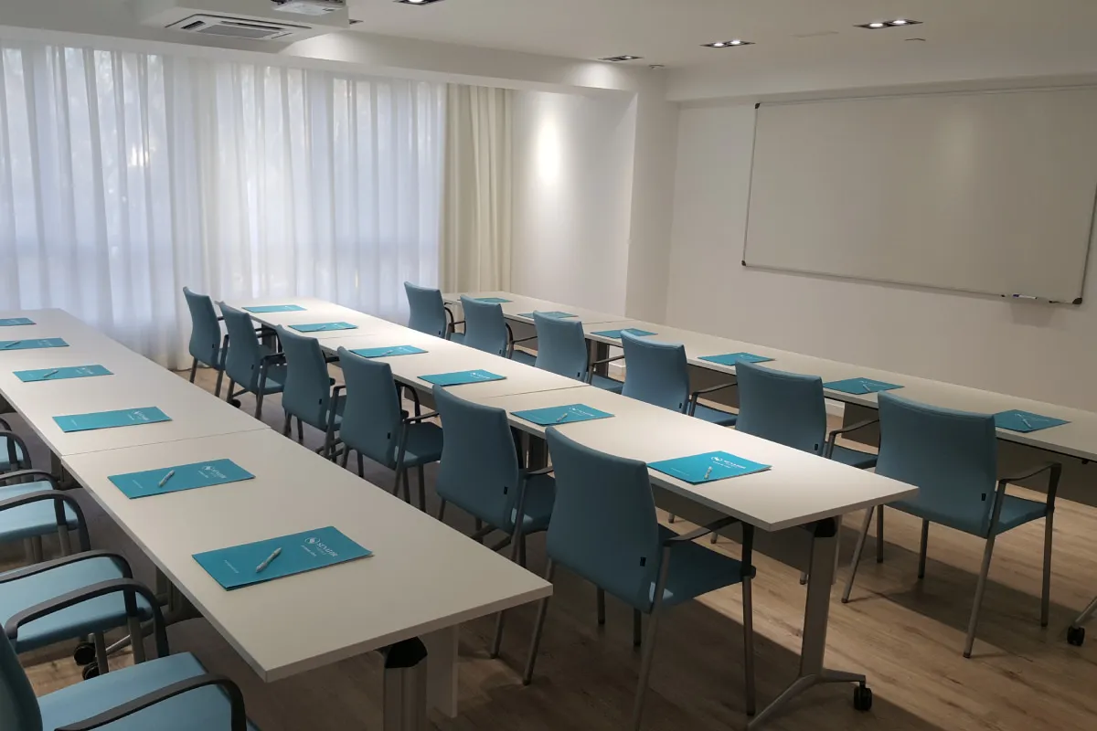 Aula con mesas rectangulares, sillas azules y proyector en el techo.