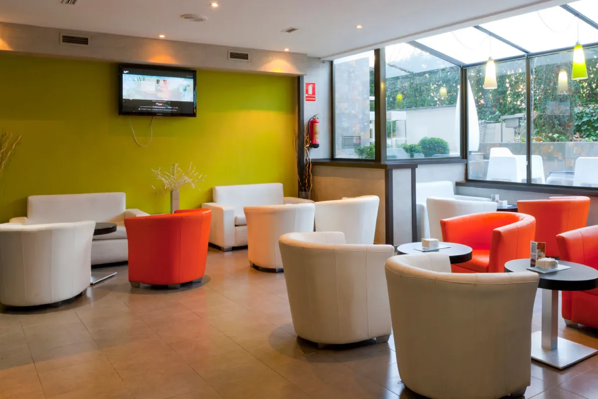 Sala de espera moderna con sof&aacute;s blancos y naranjas, televisi&oacute;n y ventanales