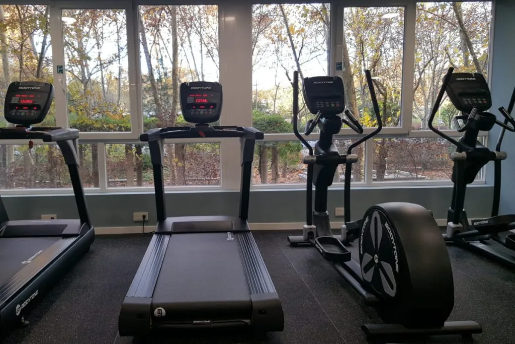 Gimnasio con cinta, elíptica y bicicleta, máquinas frente a ventanas.