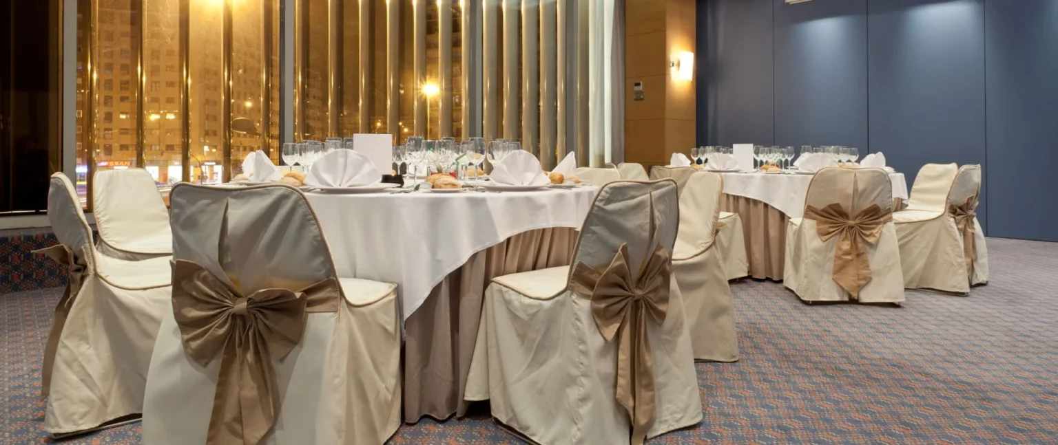 Mesa de banquete con manteles blancos y mo&ntilde;os beige, en sal&oacute;n con ventanales.
