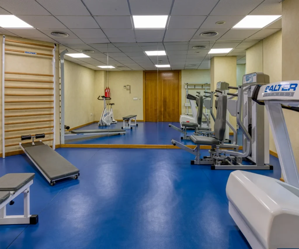 Gimnasio con m&aacute;quinas y camilla de ejercicios, suelo azul y techo de rejilla
