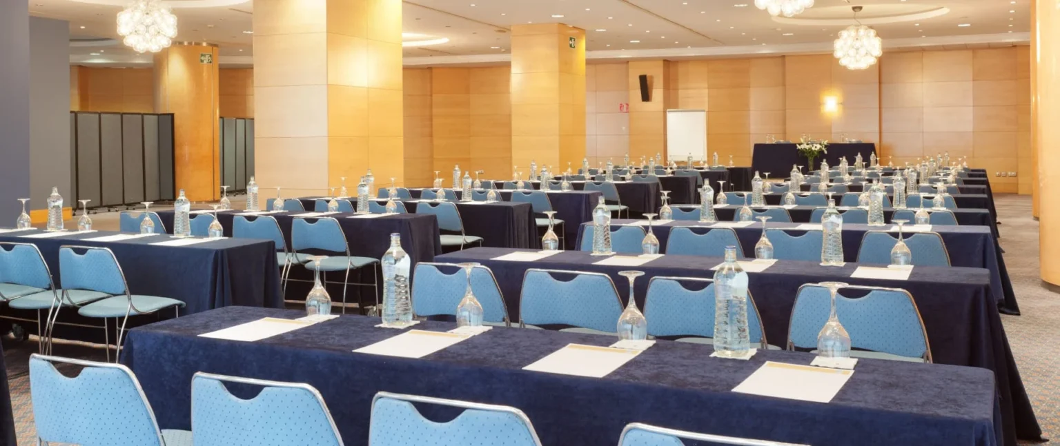 Sala de eventos con mesas largas, sillas azules y botellas sobre mantel.