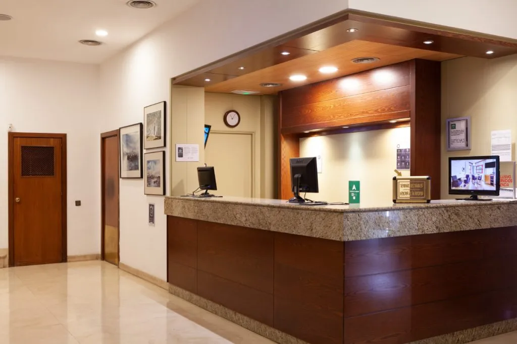 Recepción de hotel con mostrador de madera, monitores y sala de espera.
