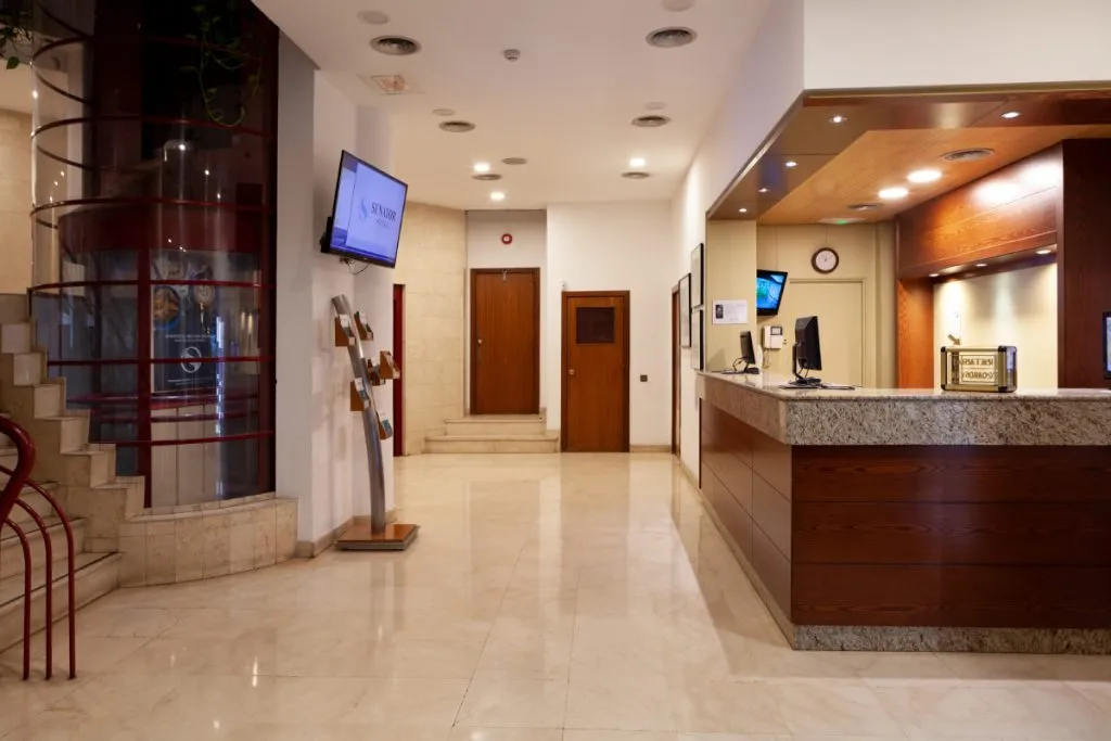 Pasillo interior de oficina con recepción, pantallas y puertas de madera.