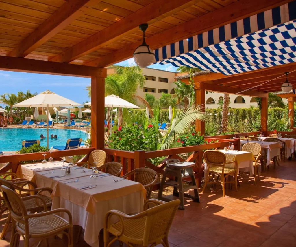 Terraza de hotel con mesas, sombrillas y piscina con plantas y toldos azules