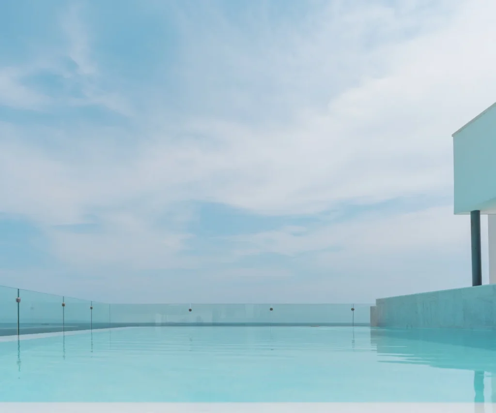 Piscina azul con barandilla de vidrio frente al mar y cielo nublado