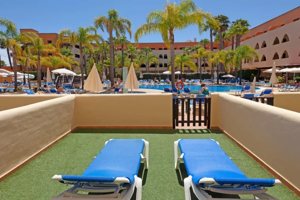 Terraza con c&eacute;sped y dos camillas azules frente a piscina y palmeras