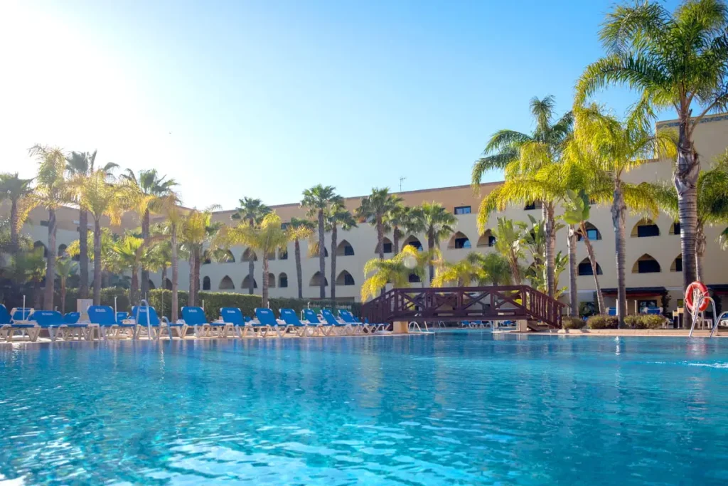 Piscina azul con palmeras y edificio blanco frente al agua, cielo despejado.