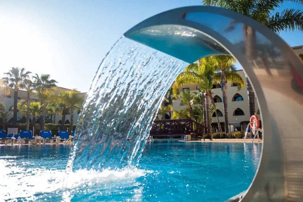 Fuente de agua y piscina con palmeras y hotel al fondo.