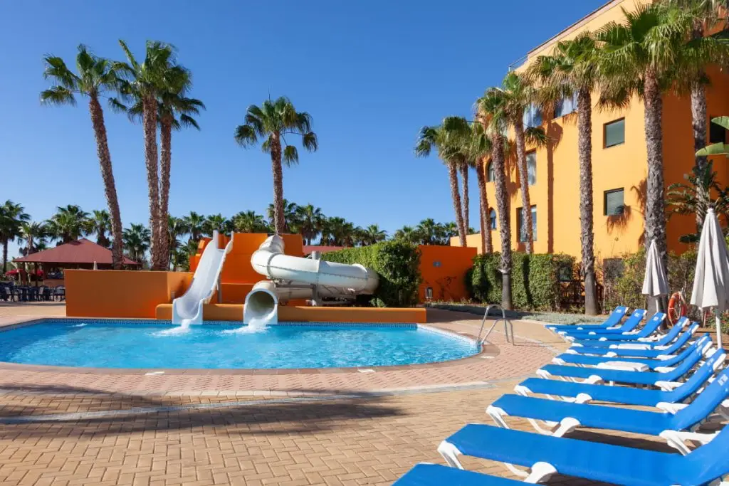 Piscina al aire libre con tobogán, palmeras y camastros azules.