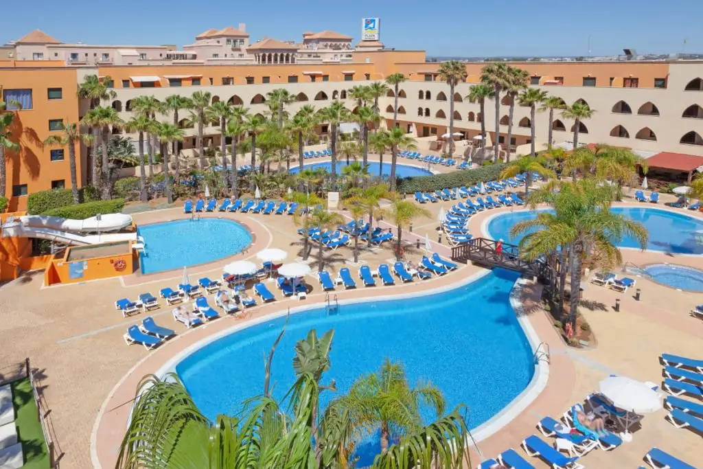 Hotel con varias piscinas azules y tumbonas, rodeado de palmeras.