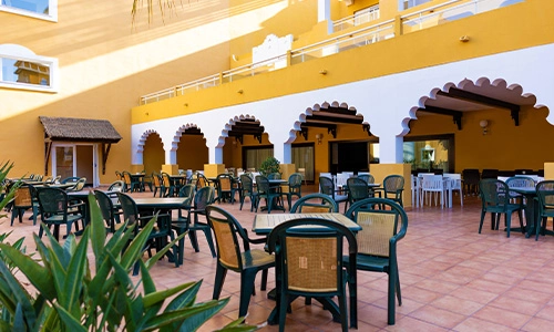 Patio con mesas y sillas de mimbre bajo arcos en edificio amarillo.